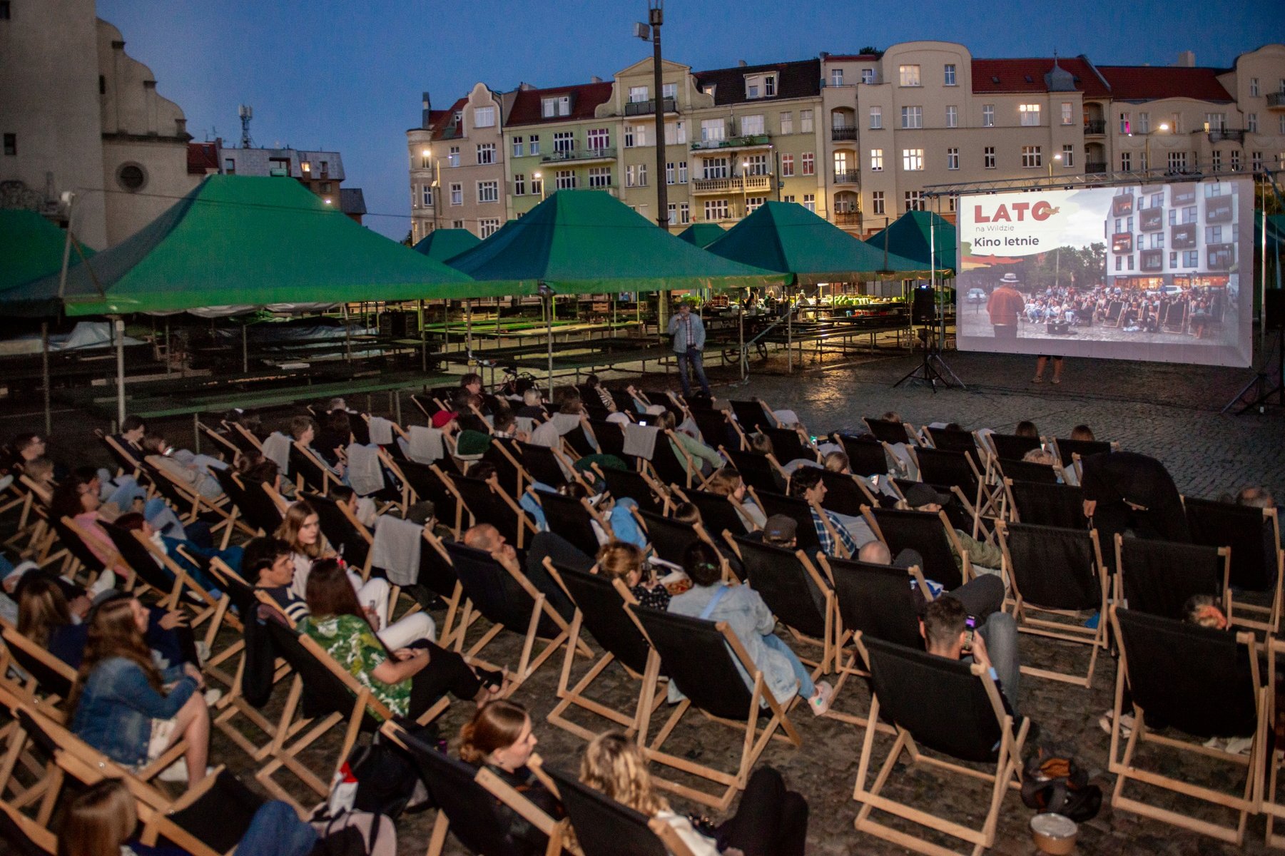 Na zdjęciu widać osoby siedzące na leżakach na rynku Wildeckim i oglądające film na ekrania - grafika rozmowy