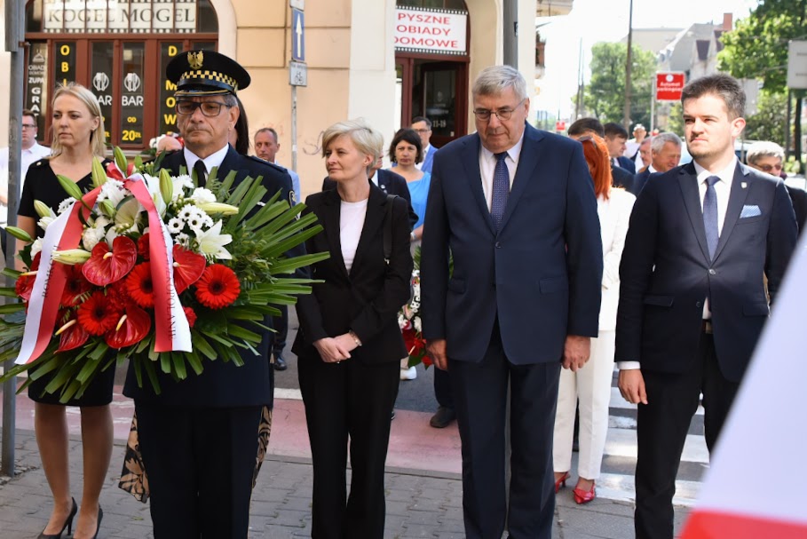 Na zdjęciu kilka osób składających wiązankę kwiatów, na pierwszym planie strażnik miejski z kwiatami - grafika rozmowy