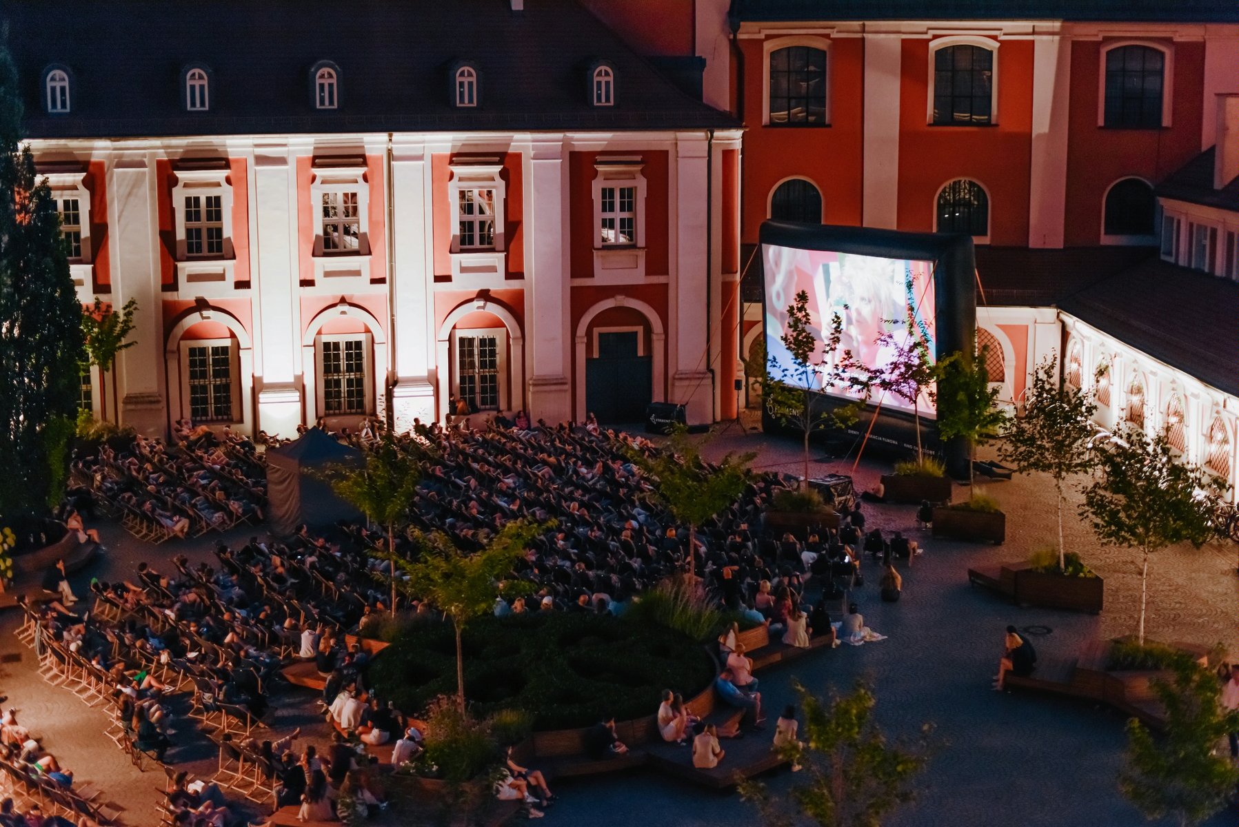 Zdjęcie przedstawia ludzi na leżakach oglądających film na telebimie na dziedzińcu urzędu miasta. - grafika rozmowy