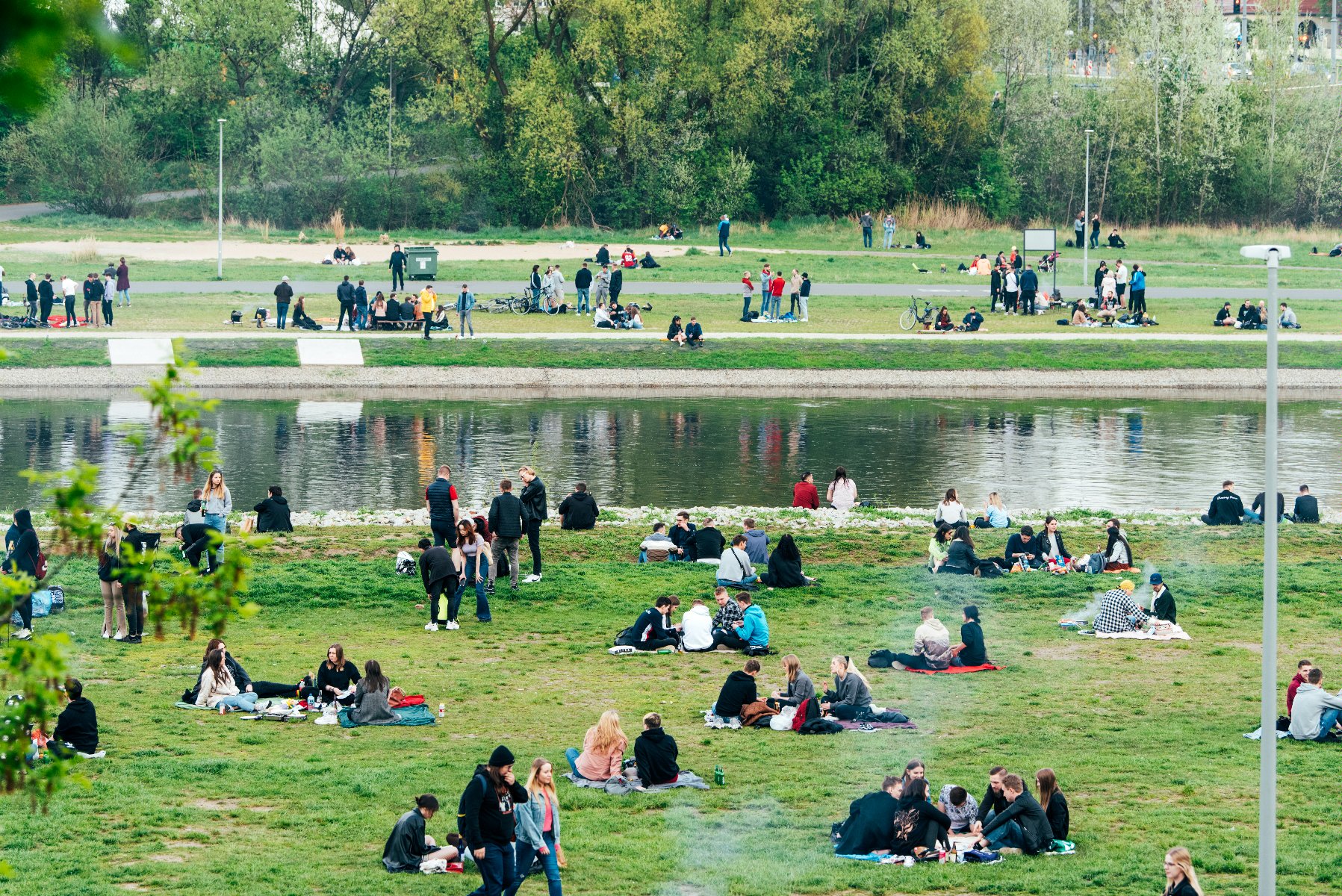 Zdjęcie przedstawia ludzi nad rzeką. Niektórzy siedzą na kocach, inni stoją i rozmawiają. - grafika rozmowy