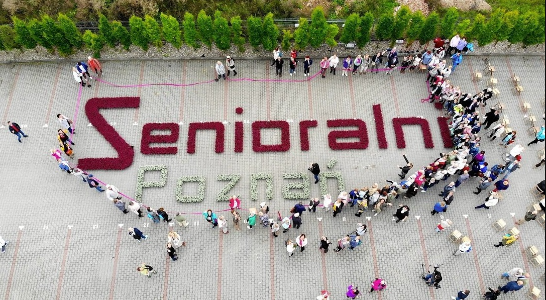 Zdjęcie z lotu ptaka, widać ludzi stojących wokół ułożonego z kwiatów napisu: Senioralni Poznań - grafika rozmowy