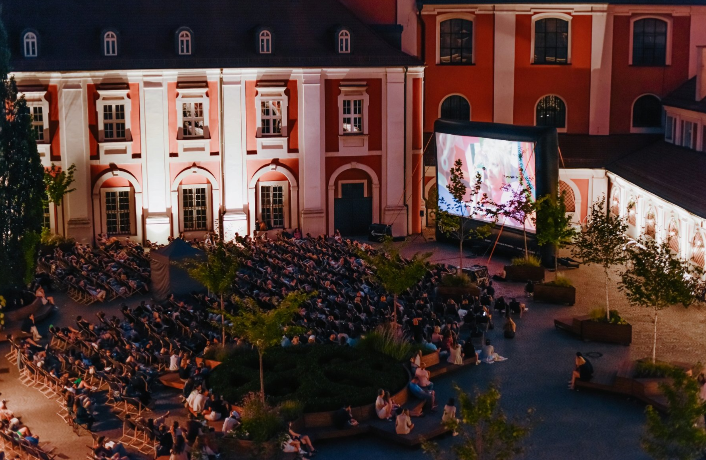 Zdjęcie przedstawia ludzi na leżakach oglądających film na telebimie na dziedzińcu urzędu miasta. - grafika rozmowy