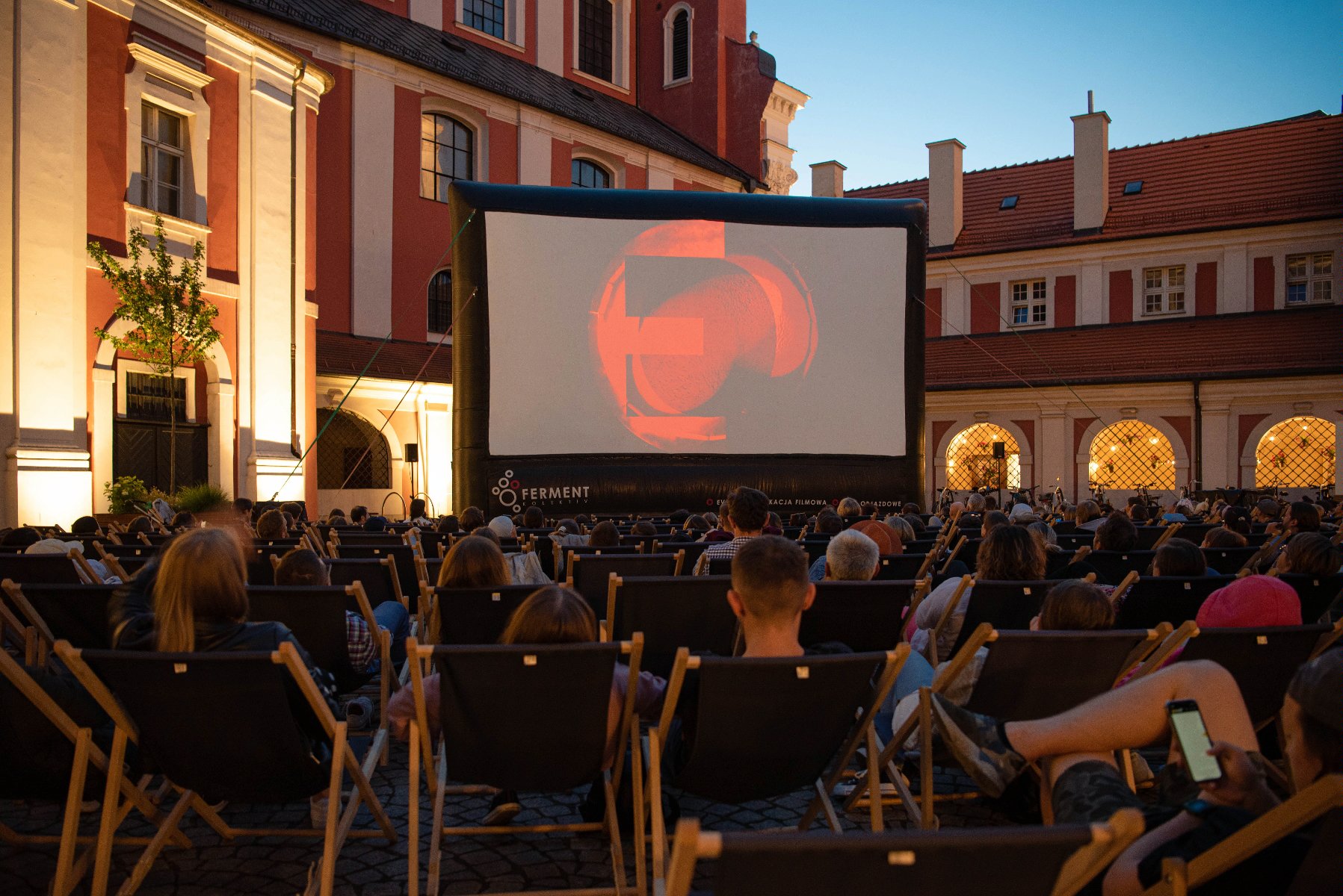 Zdjęcie przedstawia ludzi na leżakach oglądających film na telebimie na dziedzińcu urzędu miasta. - grafika rozmowy