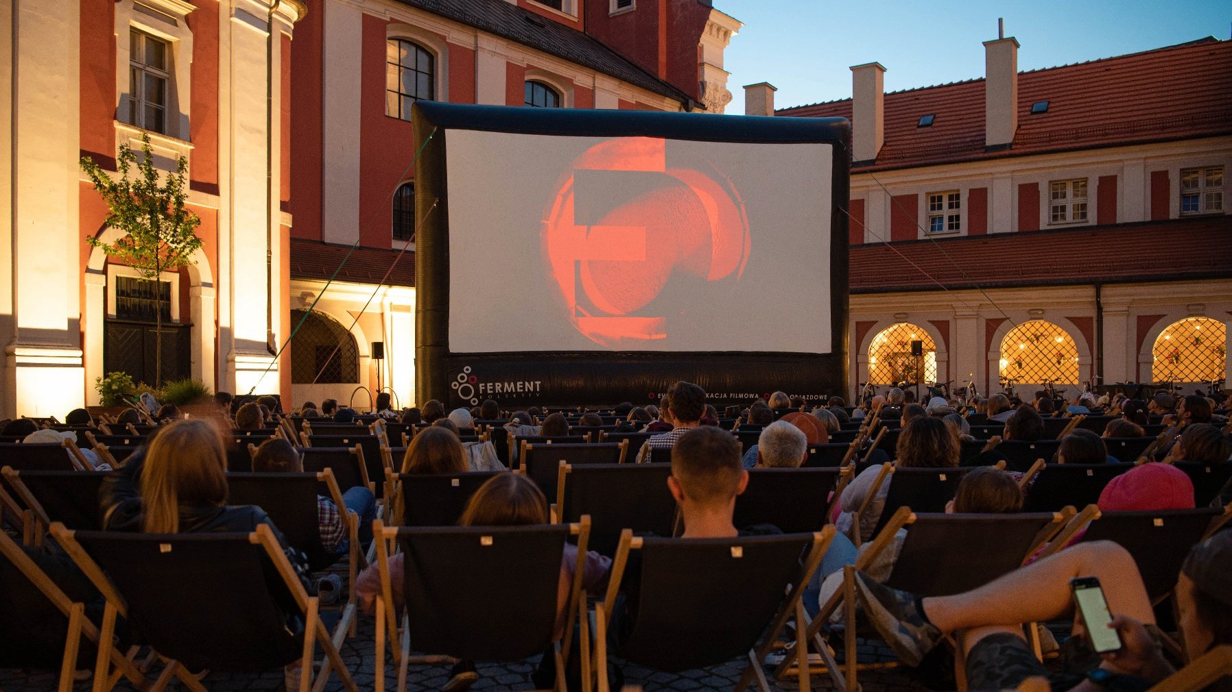 Zdjęcie przedstawia ludzi na leżakach oglądających film na telebimie na dziedzińcu urzędu miasta. - grafika rozmowy