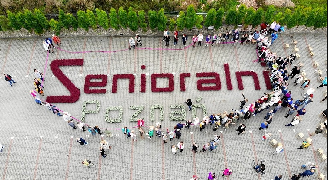 Na zdjęciu grupa ludzi wokół napisu z kwiatów: senioralni Poznań - grafika rozmowy