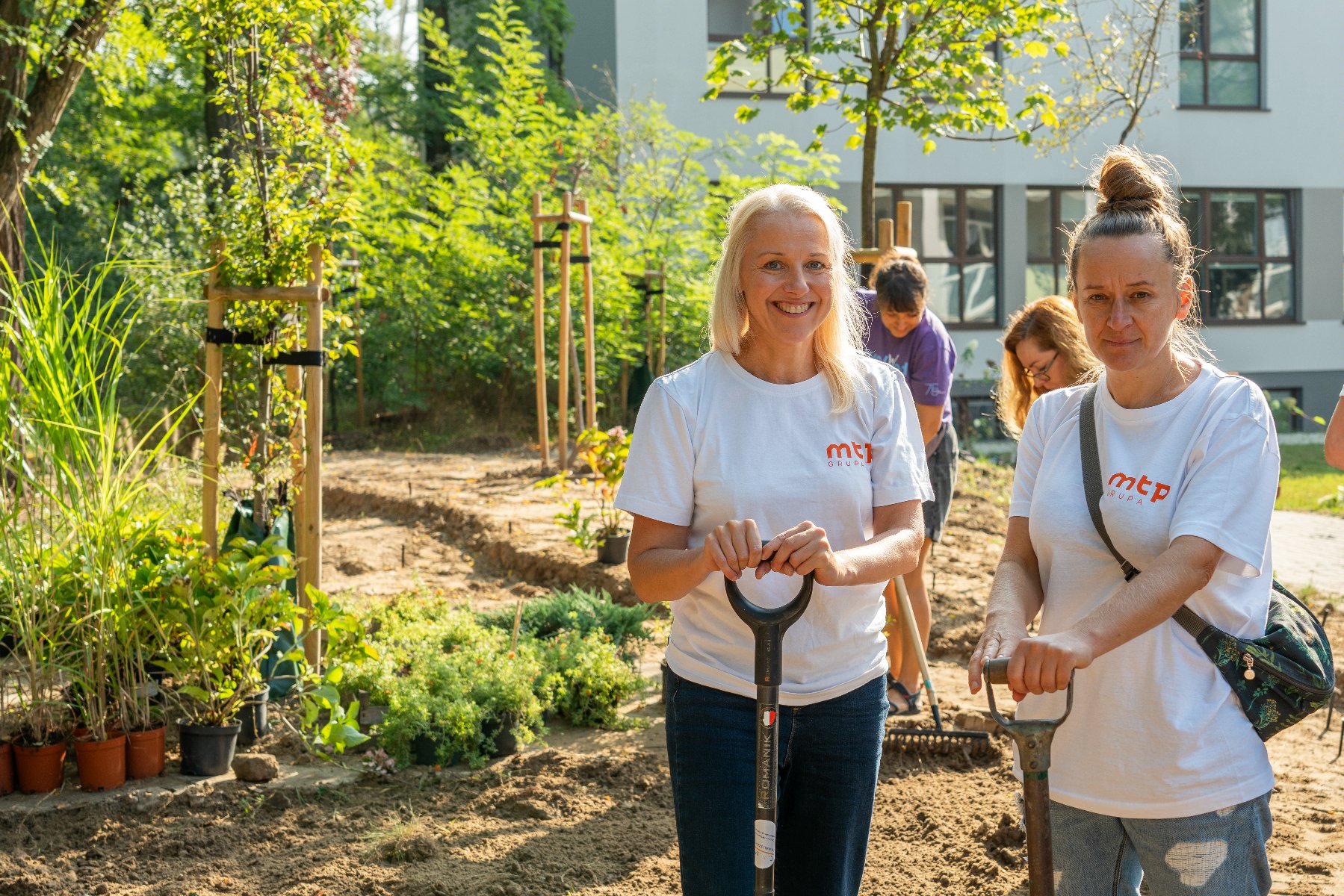 Na zdjęciu dwie uśmiechnięte wolontariuszki z łopatami, w tle drzewa - grafika rozmowy