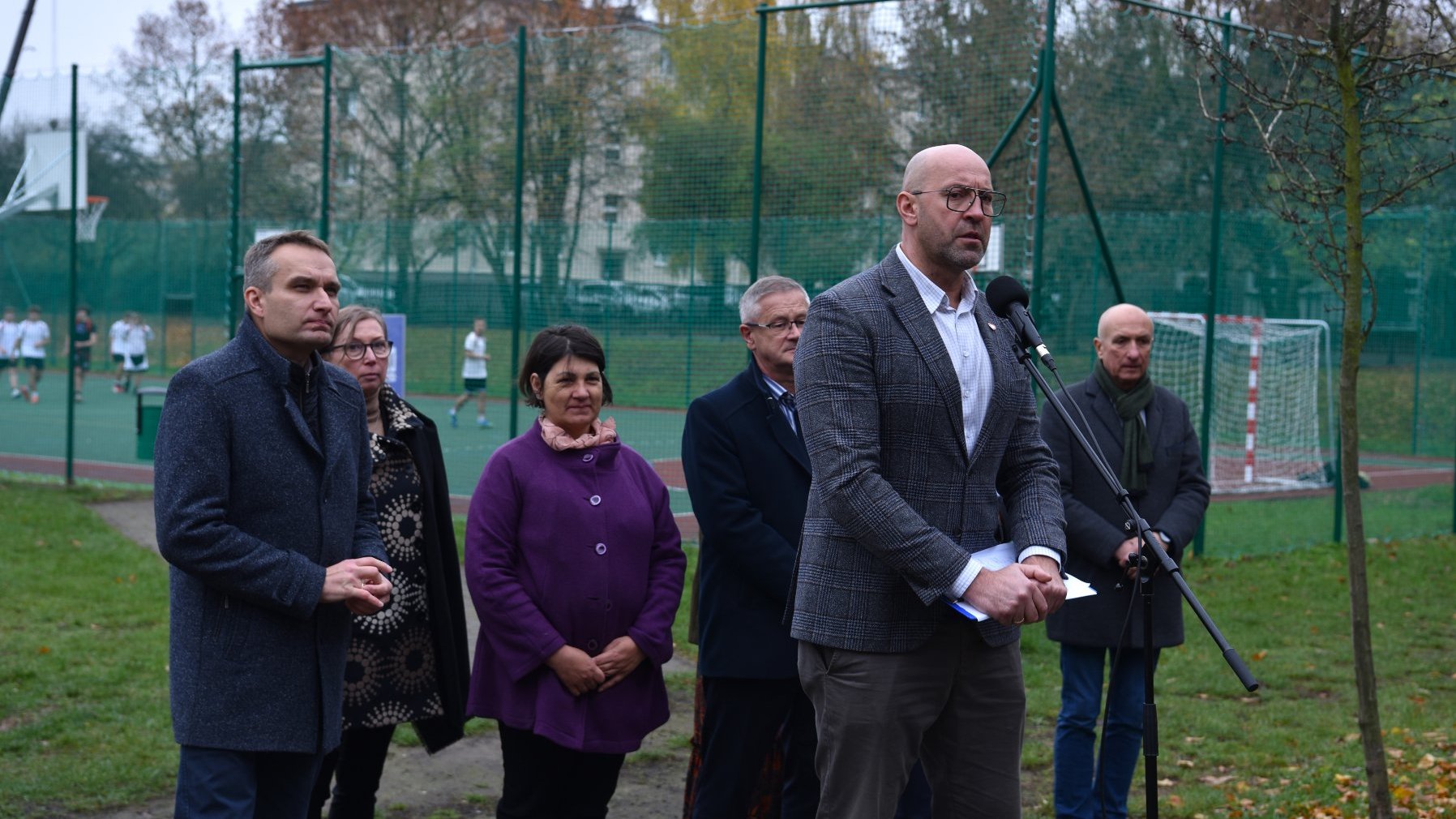 Zdjęcie przedstawia uczestników konferencji prasowej. Do mikrofonu mówi mężczyzna. Za ich plecami znajduje się boisko.