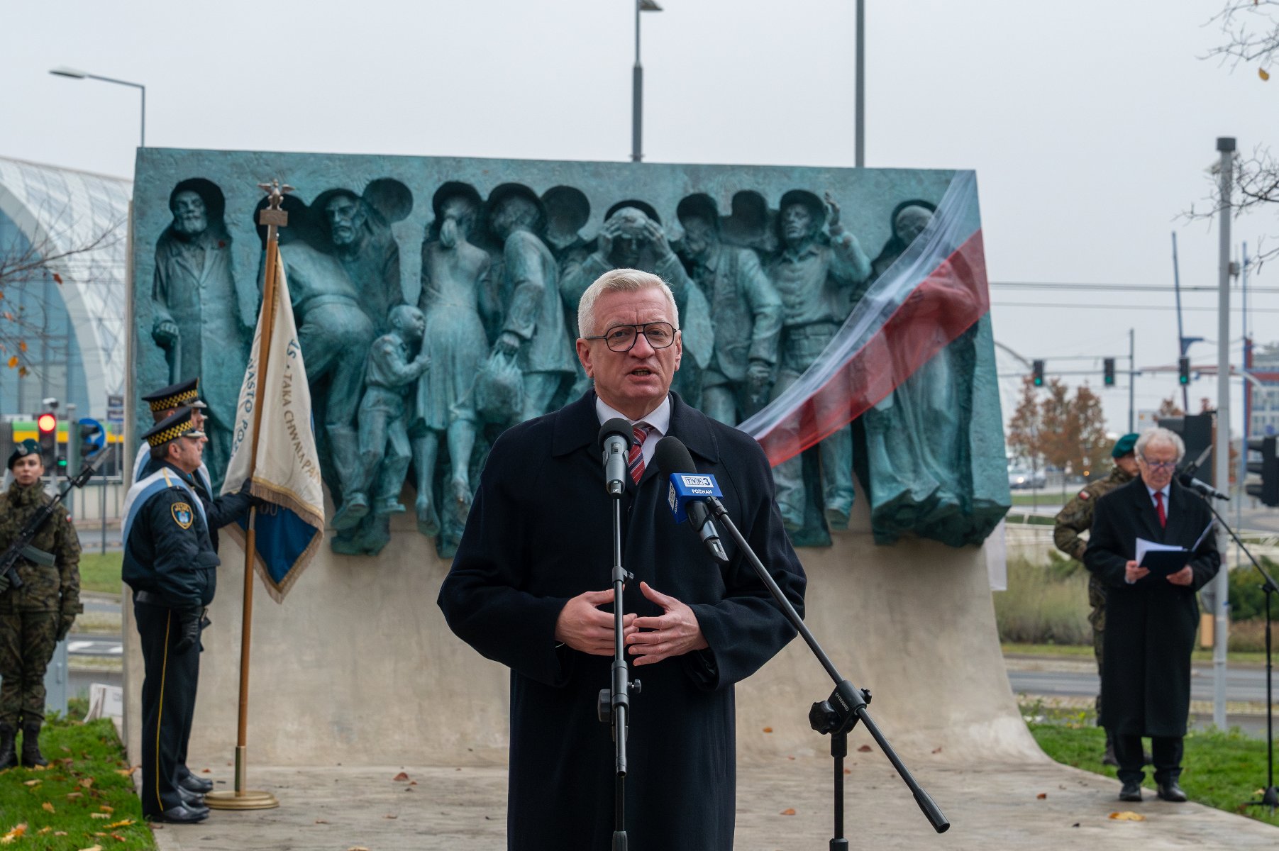 Na zdjęciu prezydent Poznania przy mikrofonie, w tle pomnik - grafika rozmowy