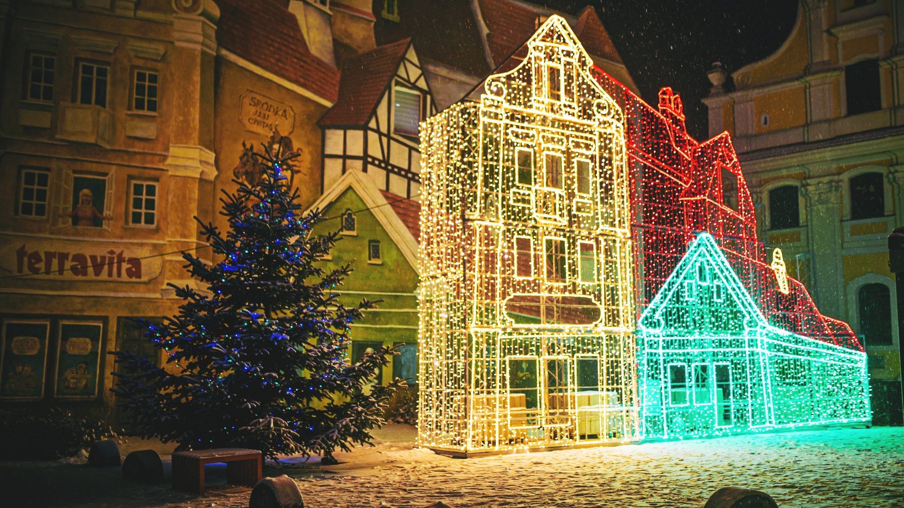 Zdjęcie przedstawia budynki. Na jednym z nich znajduje się mural. Widać też iluminację w kształcie budynków oraz ozdobioną choinkę.