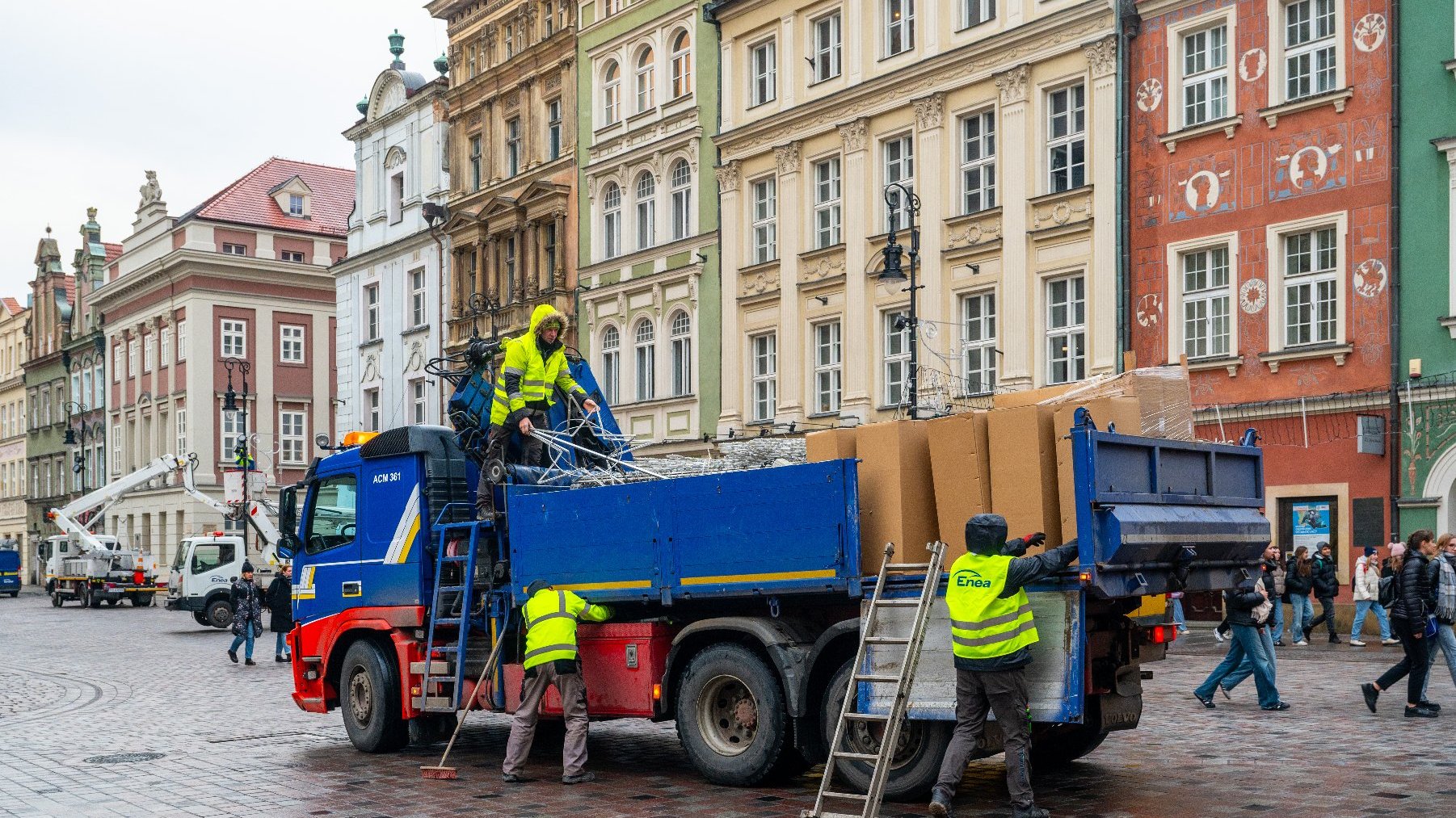 Scena na placu w europejskim mieście. Kilku pracowników w kamizelkach odblaskowych pracuje wokół dużej niebiesko-czerwonej ciężarówki załadowanej różnymi materiałami, w tym kartonowymi pudełkami i metalowymi konstrukcjami. Pracownicy zajmują się obsługą i organizowaniem tych materiałów. W tle znajdują się kolorowe, historyczne budynki z ozdobnymi detalami architektonicznymi, a także przechodnie. Na kamizelkach pracowników widnieje napis "Enea", co sugeruje nazwę firmy, dla której pracują.