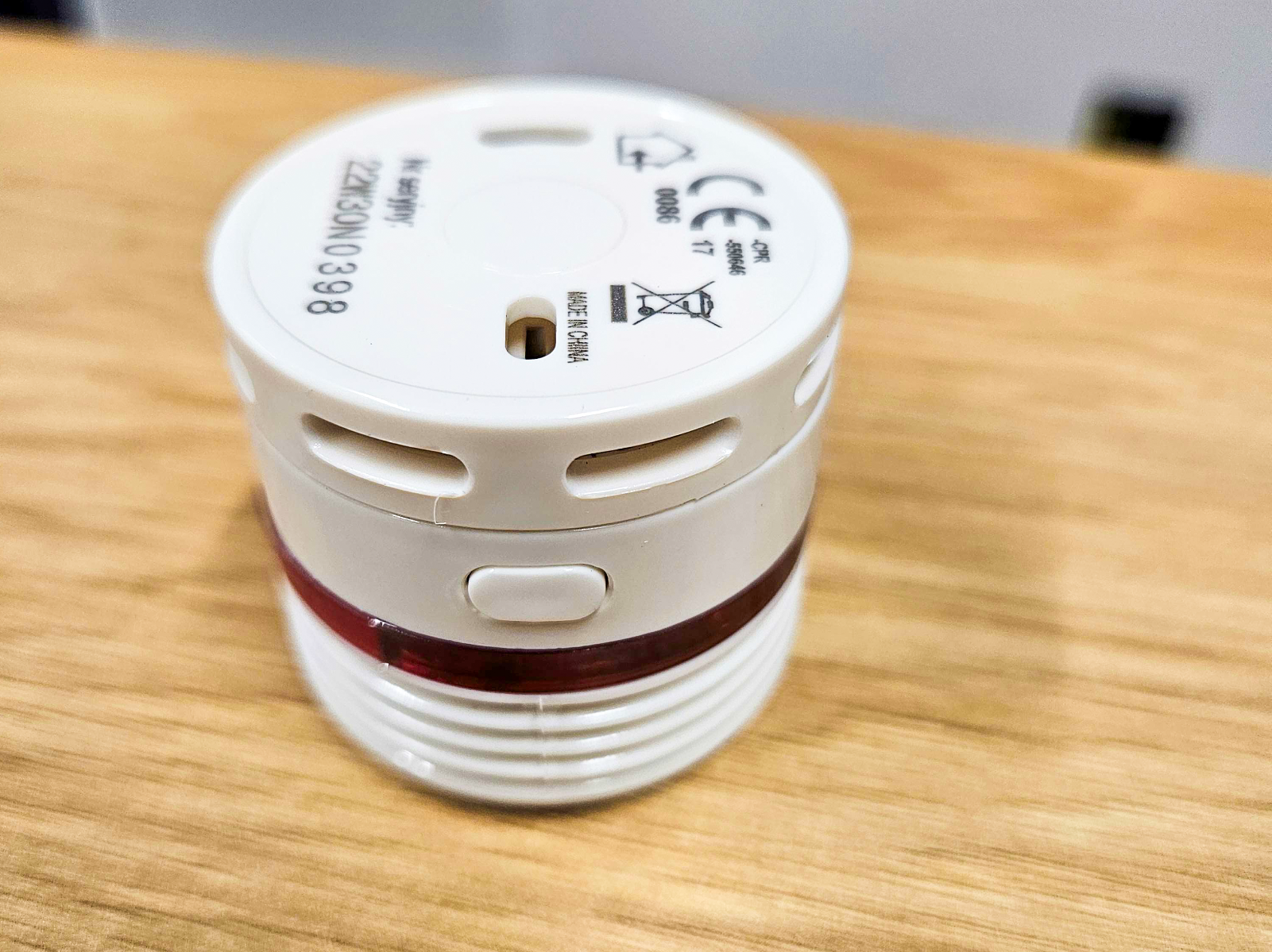 Urządzenie elektroniczne, prawdopodobnie detektor dymu lub podobne urządzenie bezpieczeństwa. Jest białe, cylindryczne z czerwoną opaską wokół środka i kilkoma otworami wentylacyjnymi po bokach. Na boku urządzenia znajduje się przycisk. Górna część urządzenia zawiera różne oznaczenia i symbole. Urządzenie leży na drewnianej powierzchni. - grafika rozmowy