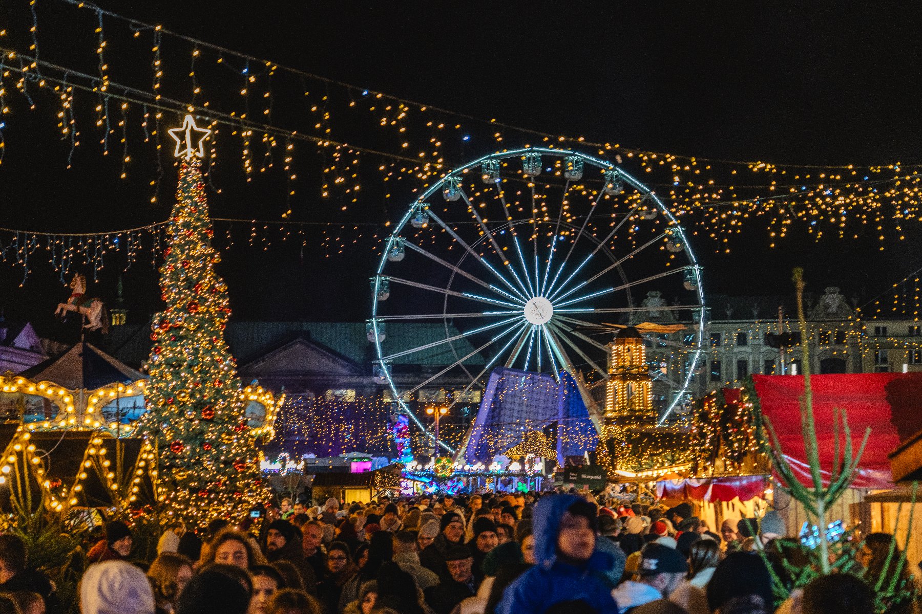 Zdjęcie przedstawia świąteczny plac Wolności. Oprócz kolorowych lampek widać na nim tłum ludzi, stoiska handlowe, choinkę i młyńskie koło. - grafika rozmowy