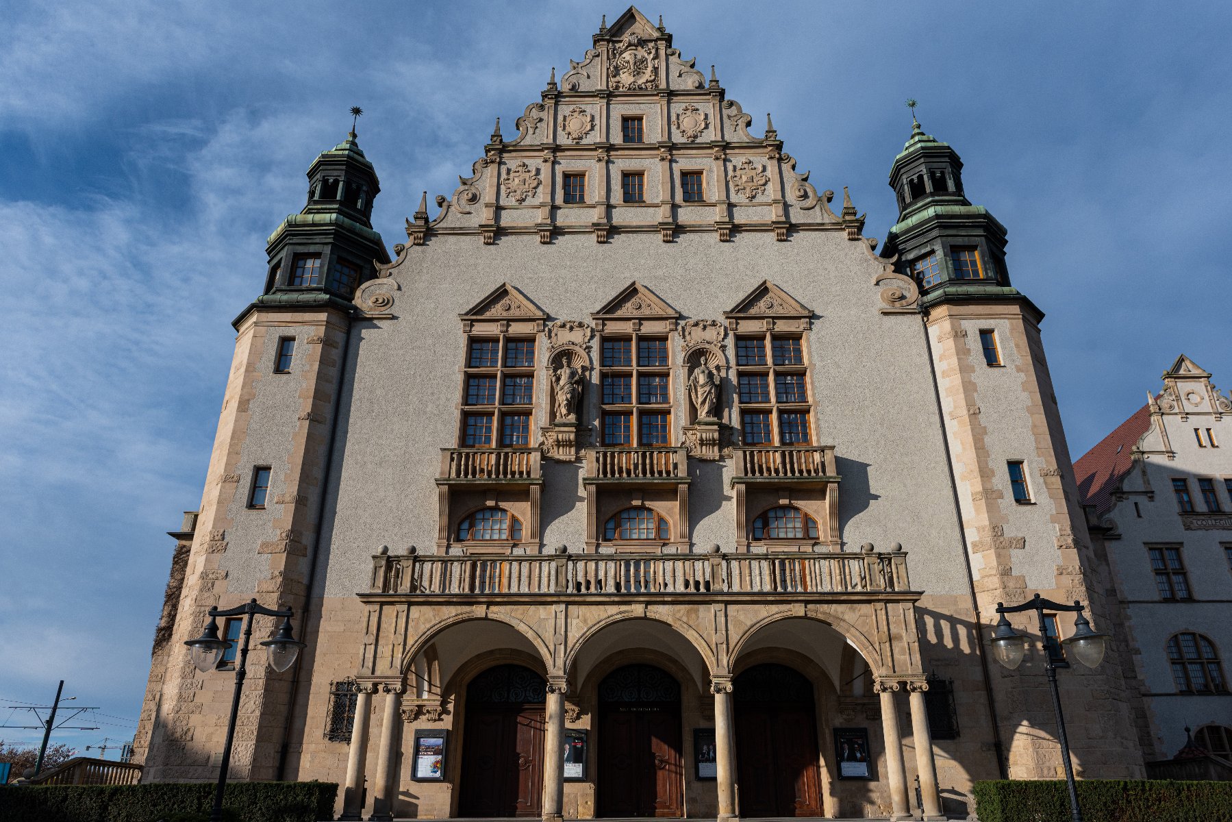 Duży ozdobny budynek o symetrycznej fasadzie. Budynek ma dwie wieże po bokach z zielonymi kopułowymi dachami. Centralna część budynku ma stromy dach z ozdobnymi elementami i posągami. Na dole znajdują się trzy łukowate wejścia, każde z drewnianymi drzwiami, a powyżej balkon. B - grafika rozmowy