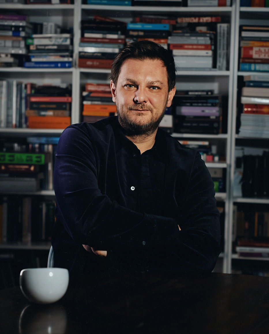 mężczyzna w cienkiej koszuli, z ciemnymi włosami i brodą na tle regału z książkami - grafika rozmowy