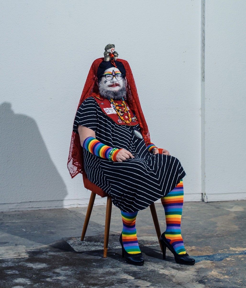 postać siedząca na krześle w tęczowym stroju i w ozdobnym nakryciu głowy, z pomalowaną twarzą - grafika rozmowy