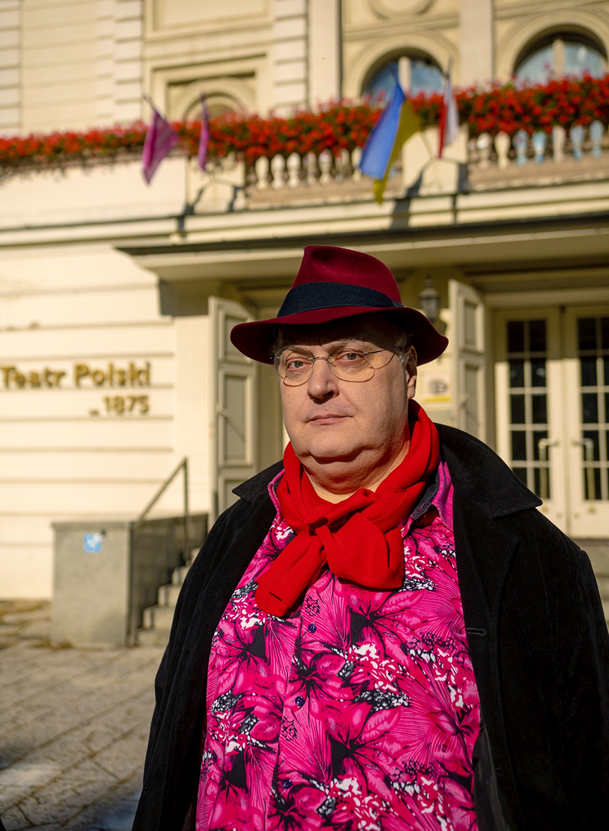 mężczyzna w kapeluszu, różowej koszuli i czerwonym szaliku na tle Teatru Polskiego w Poznaniu - grafika rozmowy