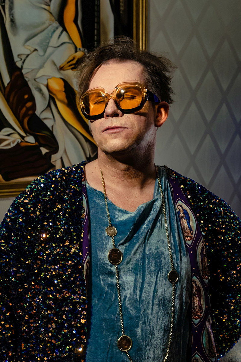 mężczyzna w dużych, pomarańczowych okularach, stroju z aksamitu i wyszywanym cekinami - grafika rozmowy