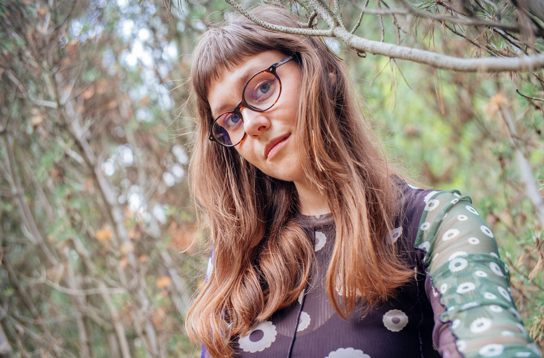 młoda kobieta z długimi włosami i w okularach na tle gałęzi drzew - grafika rozmowy