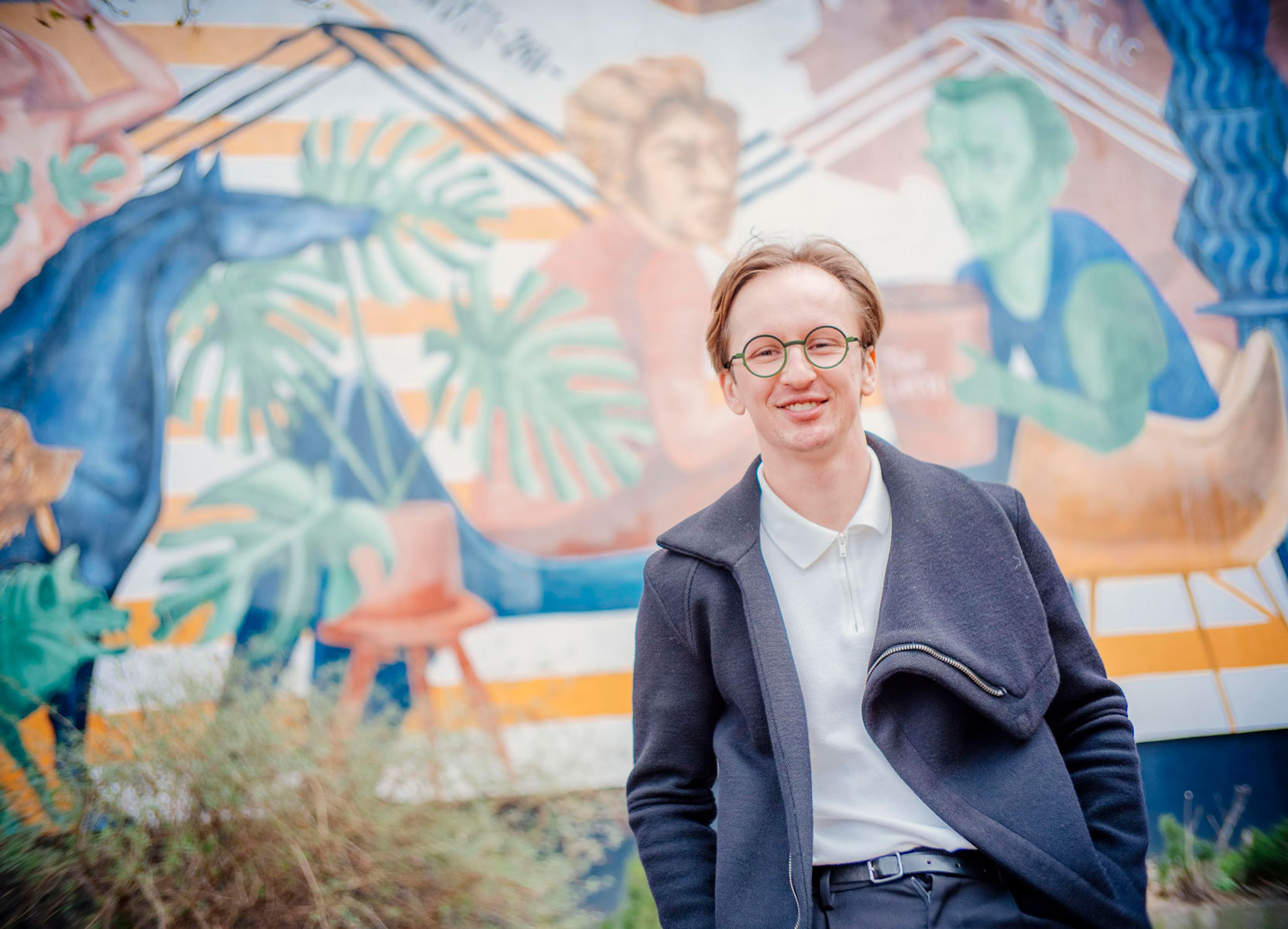 młody mężczyzna z jasnymi włosami i w okularach na tle muralu - grafika rozmowy