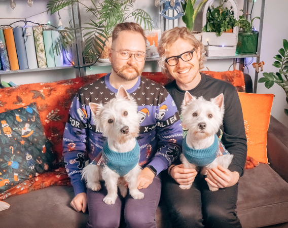 two men sitting on a couch with two dogs on their laps