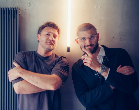 Dwóch młodych mężczyzn w krótkich włosach uśmiecha się w stronę obiektywu, jeden unosi palec wskazujący w stronę drugiego.