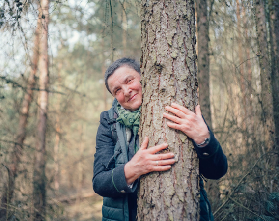 Kobieta z krótkimi, siwymi włosami na tle lasu obejmuje pień drzewa.