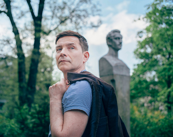 Mężczyzna z przewieszoną przez ramię marynarką stoi bokiem, patrzy w prawą stronę.