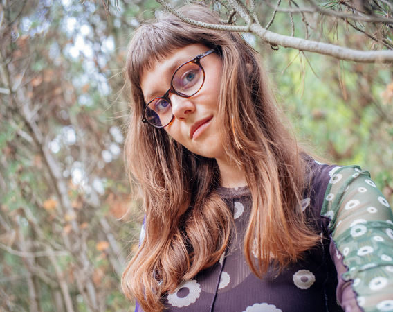 młoda kobieta z długimi włosami i w okularach na tle gałęzi drzew
