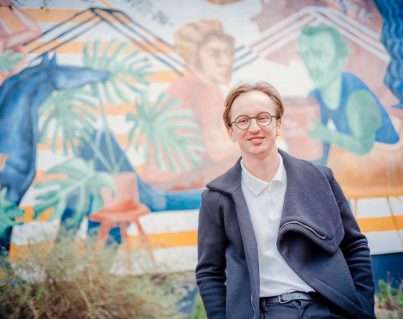 młody mężczyzna z jasnymi włosami i w okularach na tle muralu