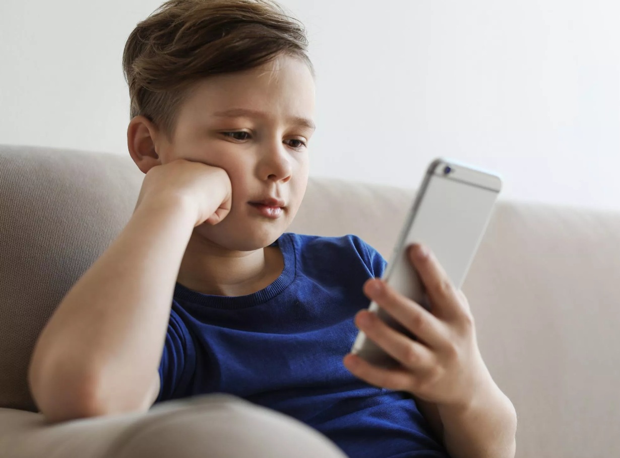 Na zdjęciu mały chłopiec wpatrujący się w ekran trzymanego w ręku smartfona