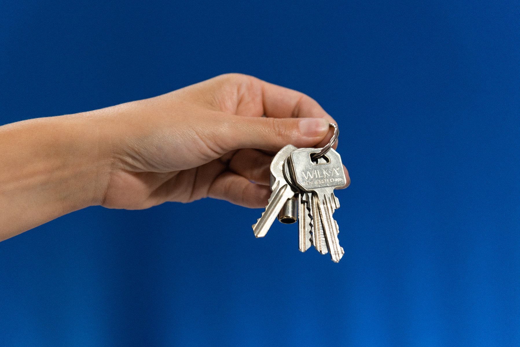 Ręka trzyma pęk metalowych kluczy na srebrnym kółku, na jednolitym niebieskim tle.