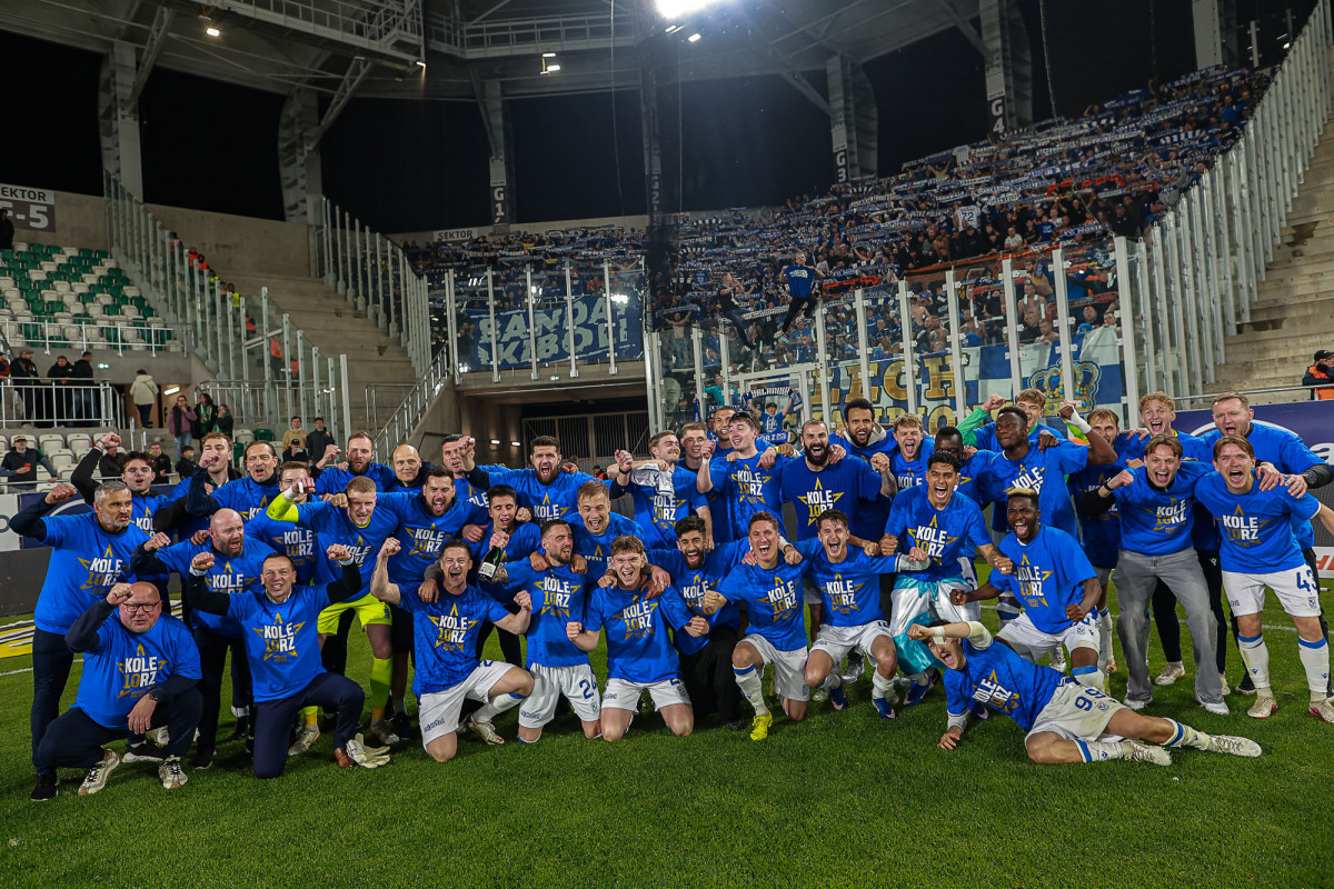 Zdjęcie przedstawia drużynę piłkarską Lecha Poznań świętującą sukces na murawie stadionu. Zawodnicy oraz sztab szkoleniowy mają na sobie niebieskie koszulki z napisem "KOLE10RZ".