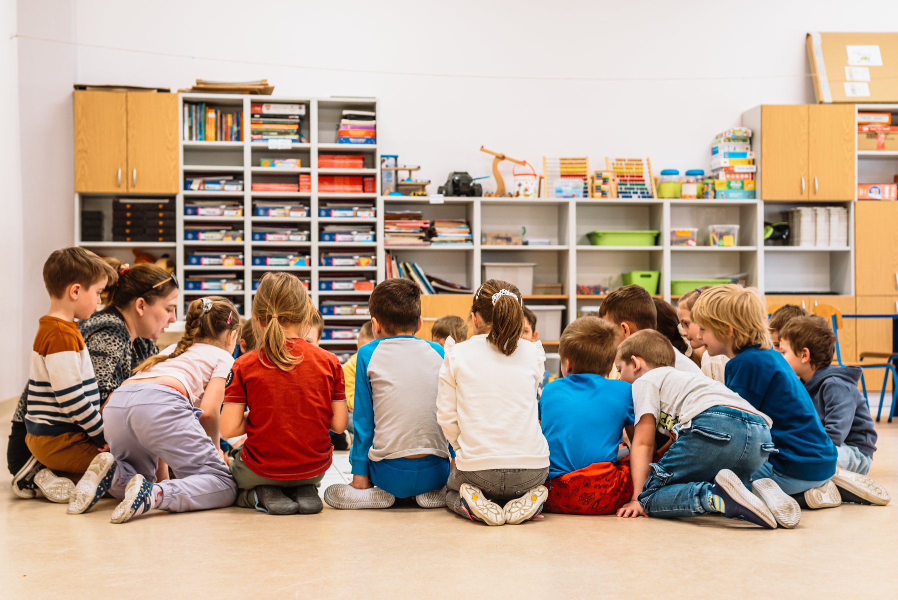 Zdjęcie przedstawia dzieci i nauczycielkę siedzących w kółku na dywanie w sali przedszkolnej. Dalej widać szafki z zabawkami.
