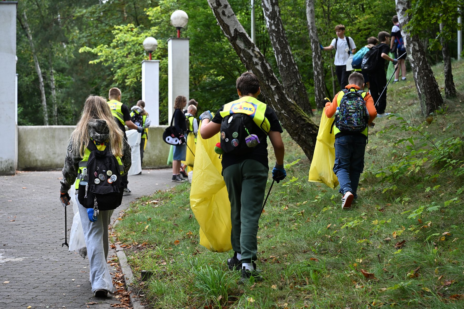 Zdjęcie przedstawia grupę młodych ludzi z workami na śmieci, chodzących wśród drzew.