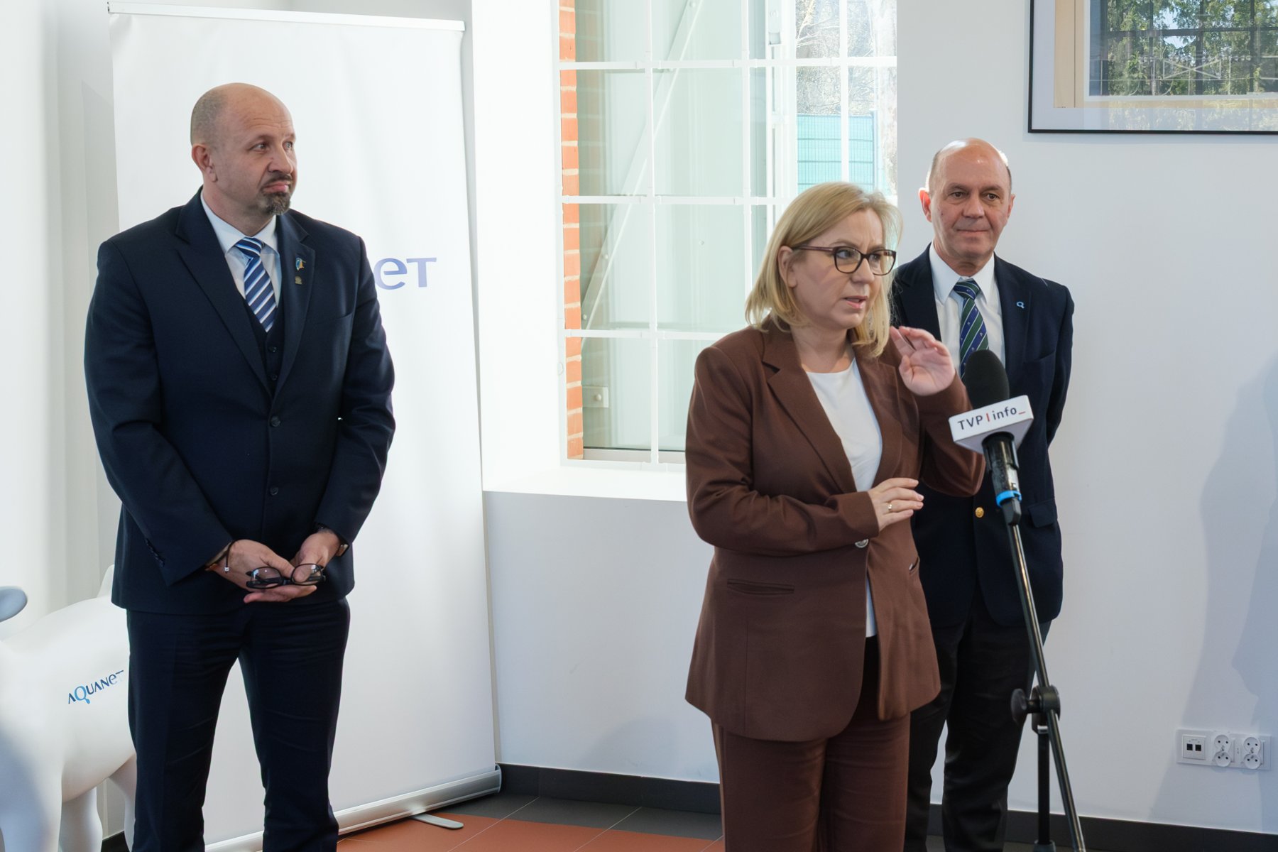 Zdjęcie przedstawia kobietę i dwóch mężczyzn stojących obok siebie. Jeden z mężczyzn stoi przed mikrofonem. Na krzesłach przed nimi siedzą ludzie.