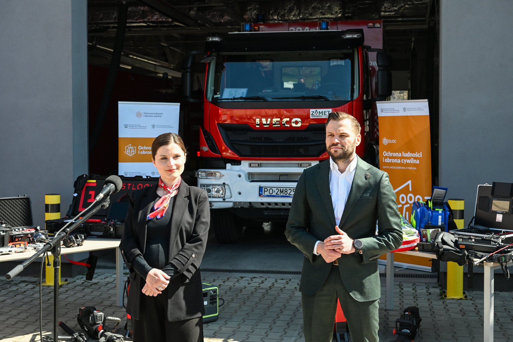 Zdjęcie przedstawia konferencję prasową lub oficjalną prezentację zorganizowaną przed garażem straży pożarnej. Na pierwszym planie stoi dwoje prelegentów: kobieta w czarnym żarcie z czerwoną apaszką oraz mężczyzna w ciemnozielonym garniturze. W tle, bezpośrednio za nimi, widoczny jest nowoczesny, czerwony wóz strażacki marki Iveco.