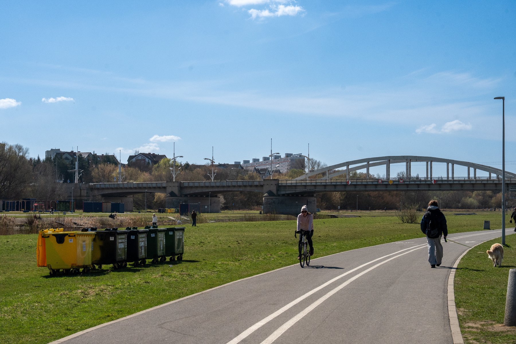 Zdjęcie przedstawia kontenery na odpady, które stoją na terenie zielonym. W oddali widać most.
