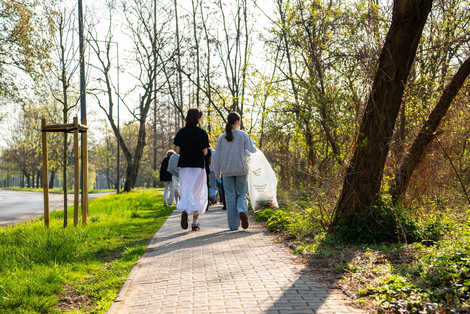 Zdjęcie przedstawia osobę idącą z workiem na śmieci. Widać tylko jej nogi. Wokół znajdują się drzewa.