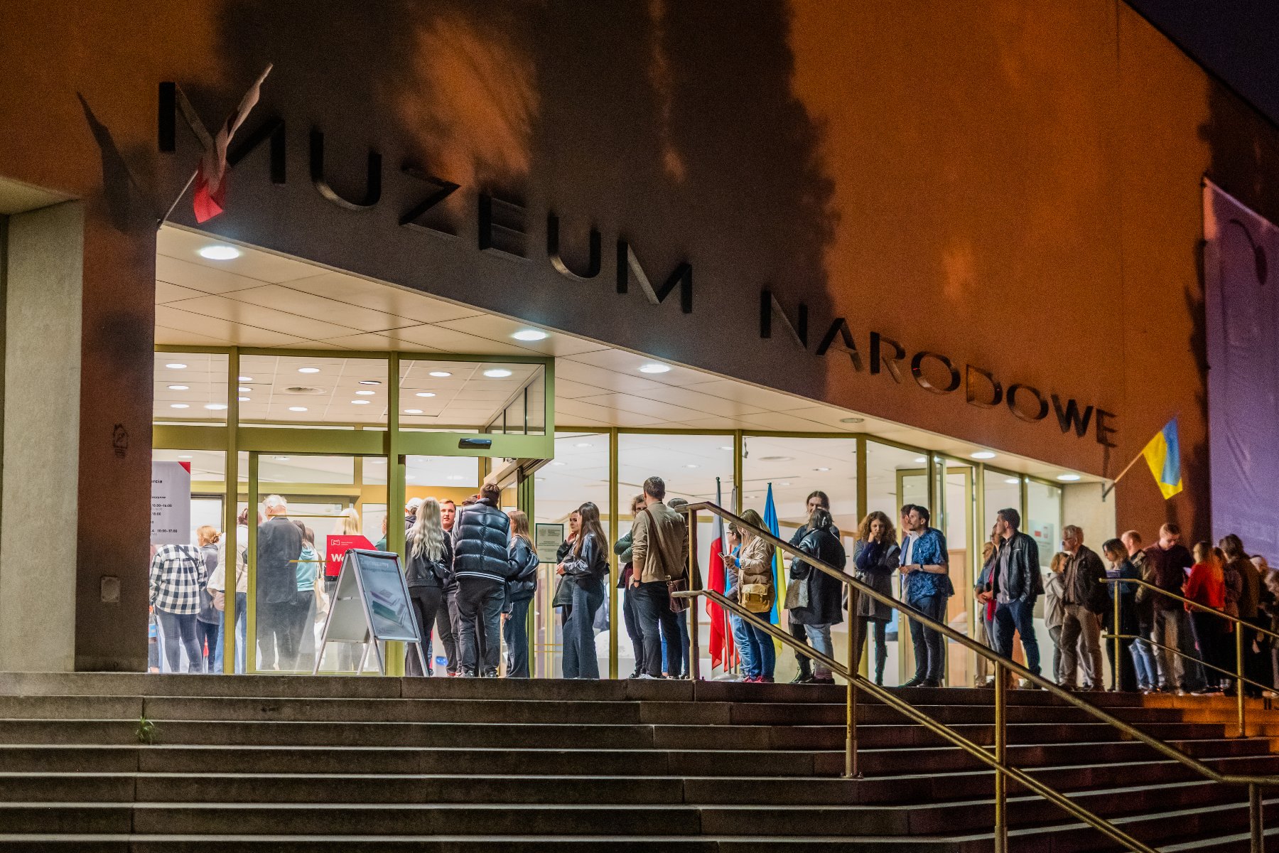 Zdjęcie przedstawia tłum ludzi w muzealnej sali. Na ścianie zawieszone są obrazy.