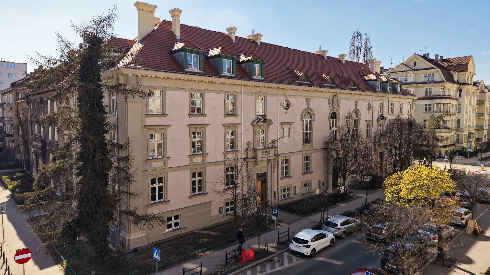 Zdjęcie przedstawia widok z góry na monumentalną kamienicę o jasnoróżowej elewacji i spadzistym, ceglanym dachu z lukarnami. Budynek posiada bogato zdobione obramowania okienne oraz dekoracyjny ryzalit z balkonem nad wejściem głównym. Przed kamienicą przebiega ulica z zaparkowanymi samochodami, a wzdłuż niej rosną drzewa, w tym jedno o wyraźnie żółtych, wiosennych liściach.