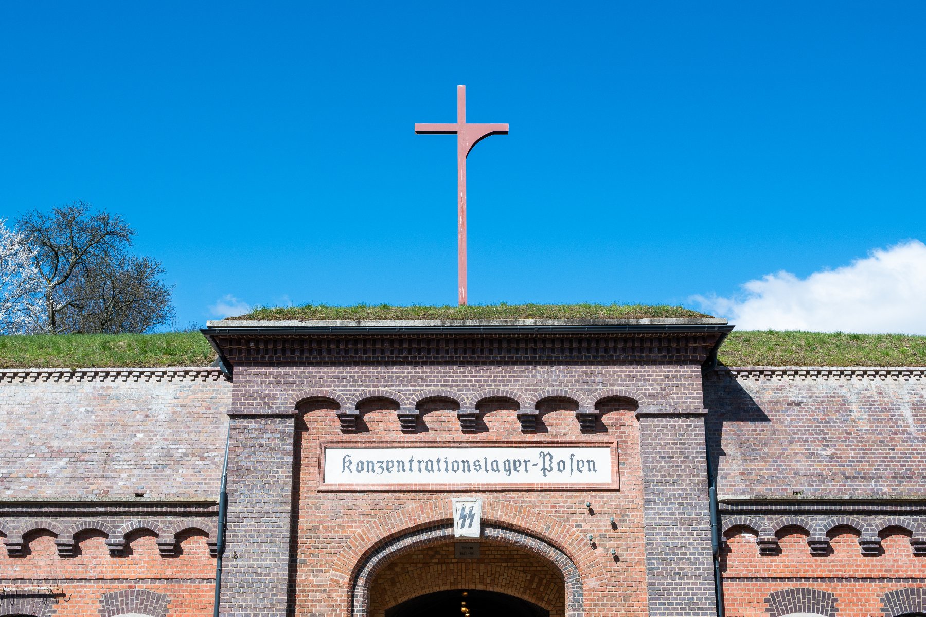 Zdjęcie przedstawia zbliżenie na górną część bramy wejściowej do historycznego obiektu z czerwonej cegły (Fort VII w Poznaniu). Nad łukiem bramy widnieje biała tablica z napisem "Konzentrationslager-Posen".